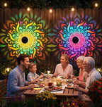 A family of five sits around a wooden dining table in a backyard at night, illuminated by warm string lights and colorful mandala-shaped solar light projections on the wooden fence behind them.