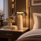 A modern table lamp with a textured beige stone base and a clear glass dome shade, glowing with warm light on a bedroom nightstand next to a bed.