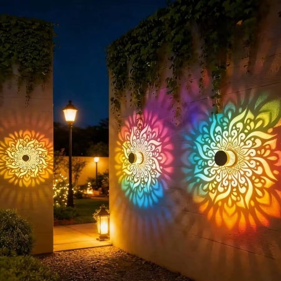 Three decorative outdoor wall lights mounted on beige walls at night, casting colorful mandala-like shadows on the surrounding walls and ground. The lights glow in pink, blue, and yellow-orange tones, with a garden path and lit lamp post visible in the background.