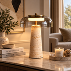 A modern table lamp with a textured beige travertine cone base, a clear glass dome shade, and a brass-toned top, lit on a marble console table in a warm, upscale living room setting.