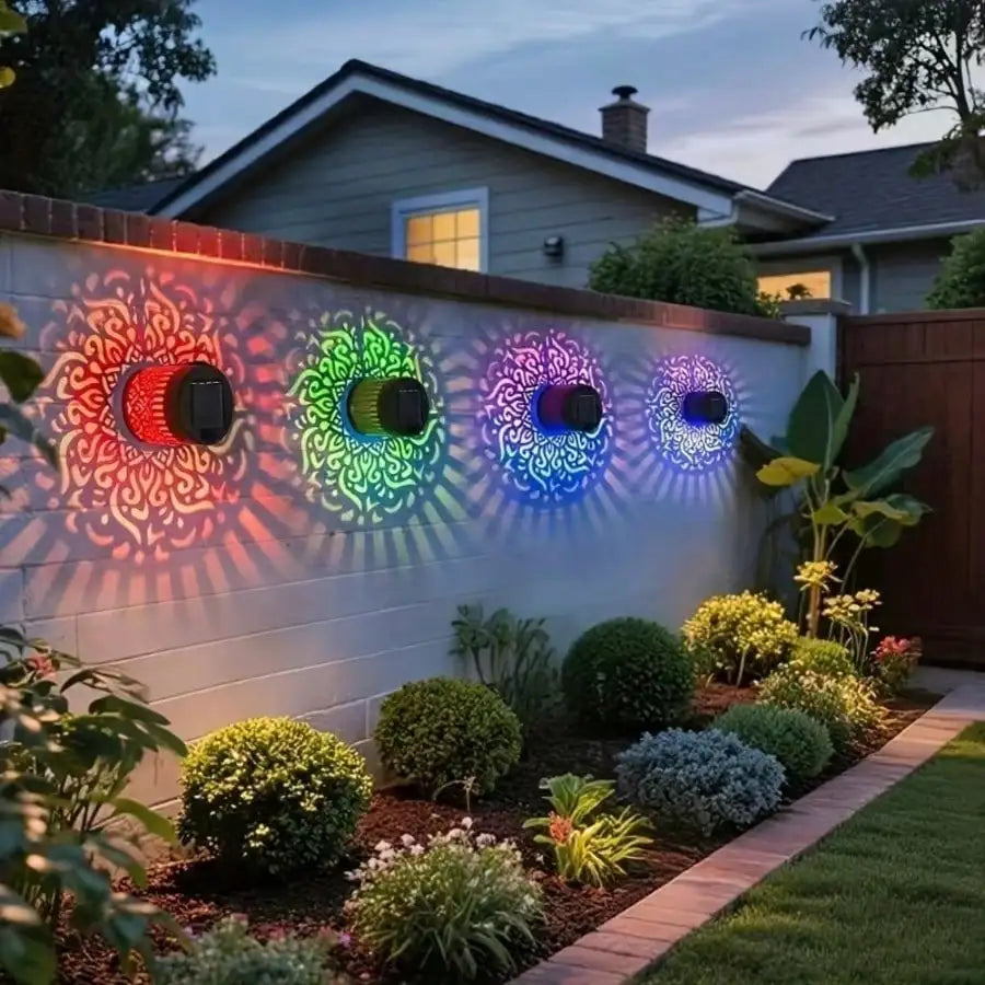 Four decorative solar wall lights mounted on a white fence in a landscaped backyard at dusk, casting colorful mandala-style shadows on the wall. The lights emit red, orange, green, blue, and purple light, with a house and garden visible in the background.