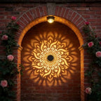 A decorative outdoor wall light mounted above an arched brick doorway, casting a warm glowing mandala shadow pattern on the brick wall. Pink roses climb the sides of the archway.