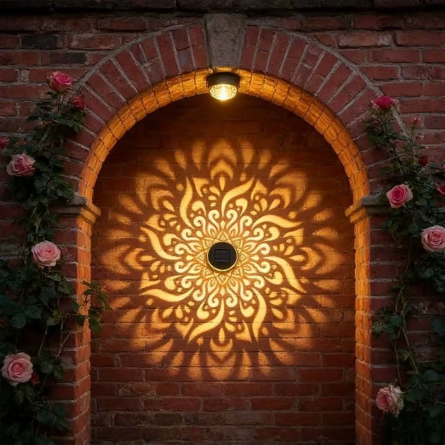 A decorative outdoor wall light mounted above an arched brick doorway, casting a warm glowing mandala shadow pattern on the brick wall. Pink roses climb the sides of the archway.