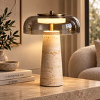 A modern table lamp with a textured beige travertine stone base, a gold-finished stem, and a clear glass dome shade, shown lit on a matching stone tabletop in a warm, styled interior setting.