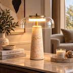 A modern table lamp with a textured beige stone base and a clear glass dome shade, illuminated with a warm light. The lamp is styled on a marble tabletop in a cozy living room setting next to a candle and books.