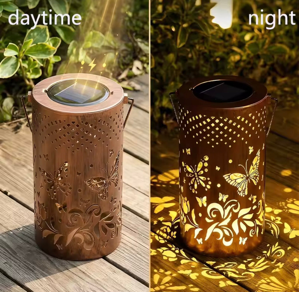 A split comparison showing a solar-powered decorative lantern in daylight on the left and at night on the right. The lantern has a coppery brown finish with laser-cut butterfly and floral patterns, featuring a built-in solar panel on top and side handles, casting a warm yellow light onto a wooden deck.