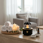 A black umbrella-shaped humidifier with misting nozzles sits on a light wood table, emitting white steam. A hand holds a white wireless remote control in the lower left, and a small bottle is on the right.