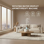 A tabletop aromatherapy water diffuser with a white base, silver umbrella-shaped top, and visible mist rising from the center. Text on the right reads 'ROTATING WATER DROPLET AROMATHERAPY MACHINE' with feature callouts including 'Simulate raindrops,' 'Colorful lights,' and 'Essential oil.